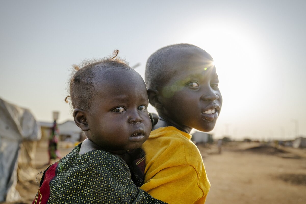 Two young children wearing green and yellow look into the camera as the sun flares behind their heads.
