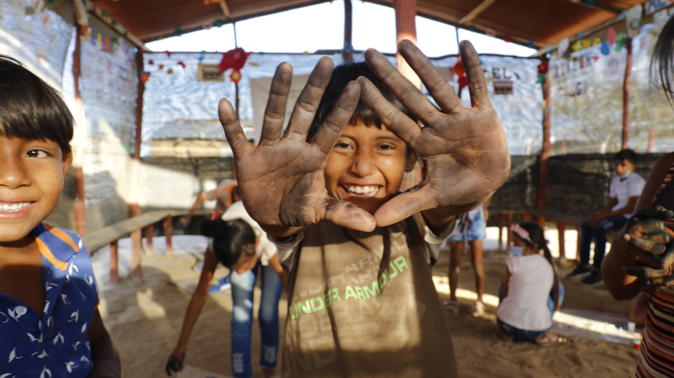 A young boy smiles and shows his dirty palms to the camera inside a tent with a dirt floor.