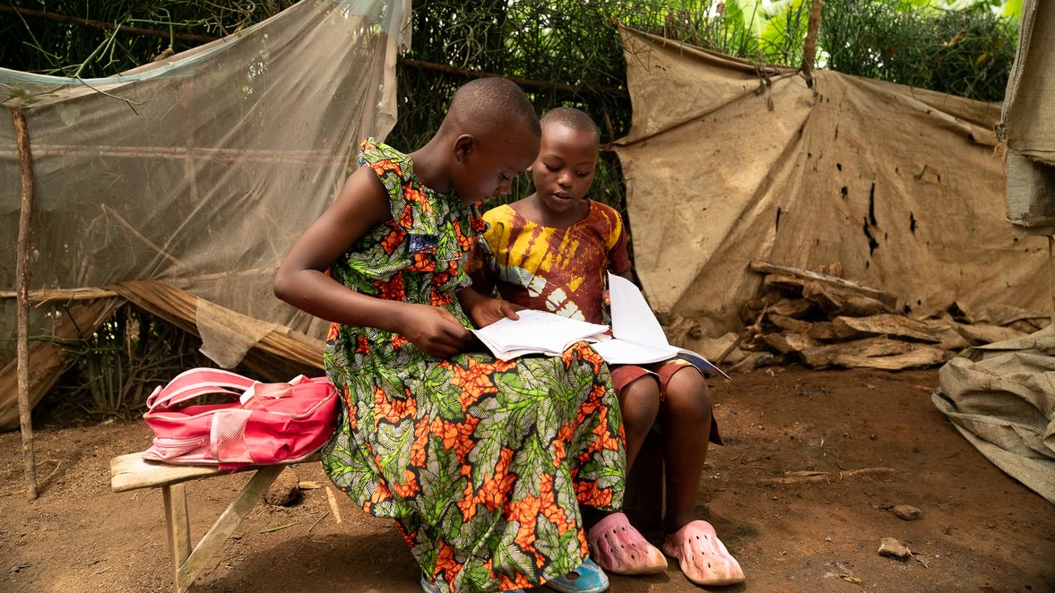 Two children sit outside in front of torn, dusty sheets, looking at a book together.