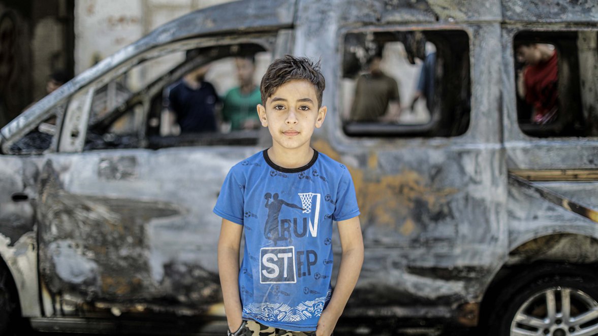 Child from Gaza, standing in front of a dishoveled car